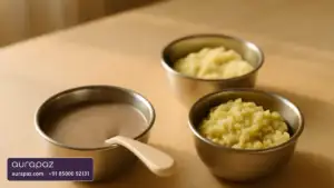 Three small steel bowls of baby first foods on a wooden table: thin ragi porridge with a baby spoon and two soft mashes